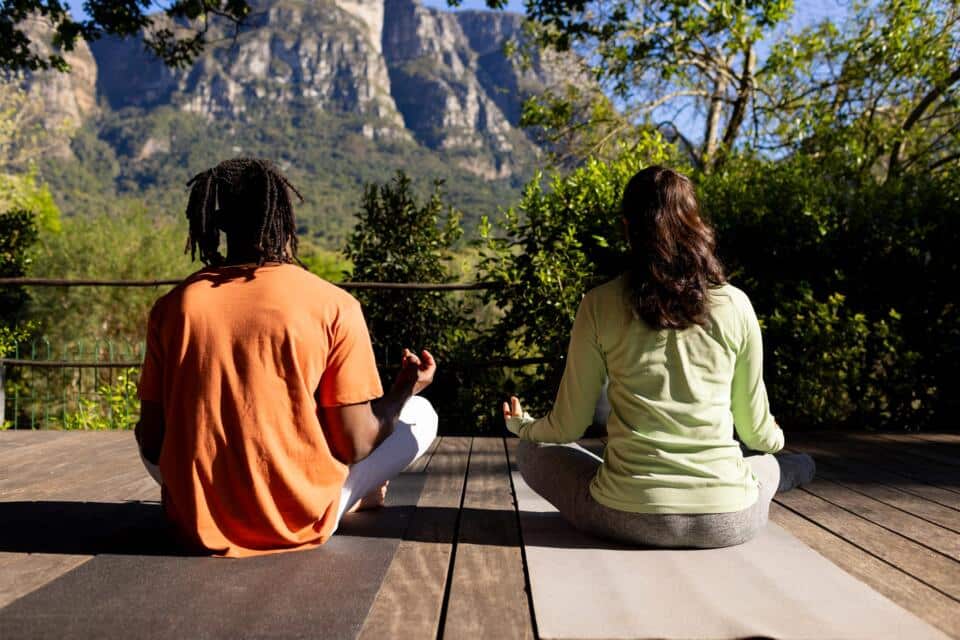 Dos personas meditando en una terraza de madera frente a una montaña