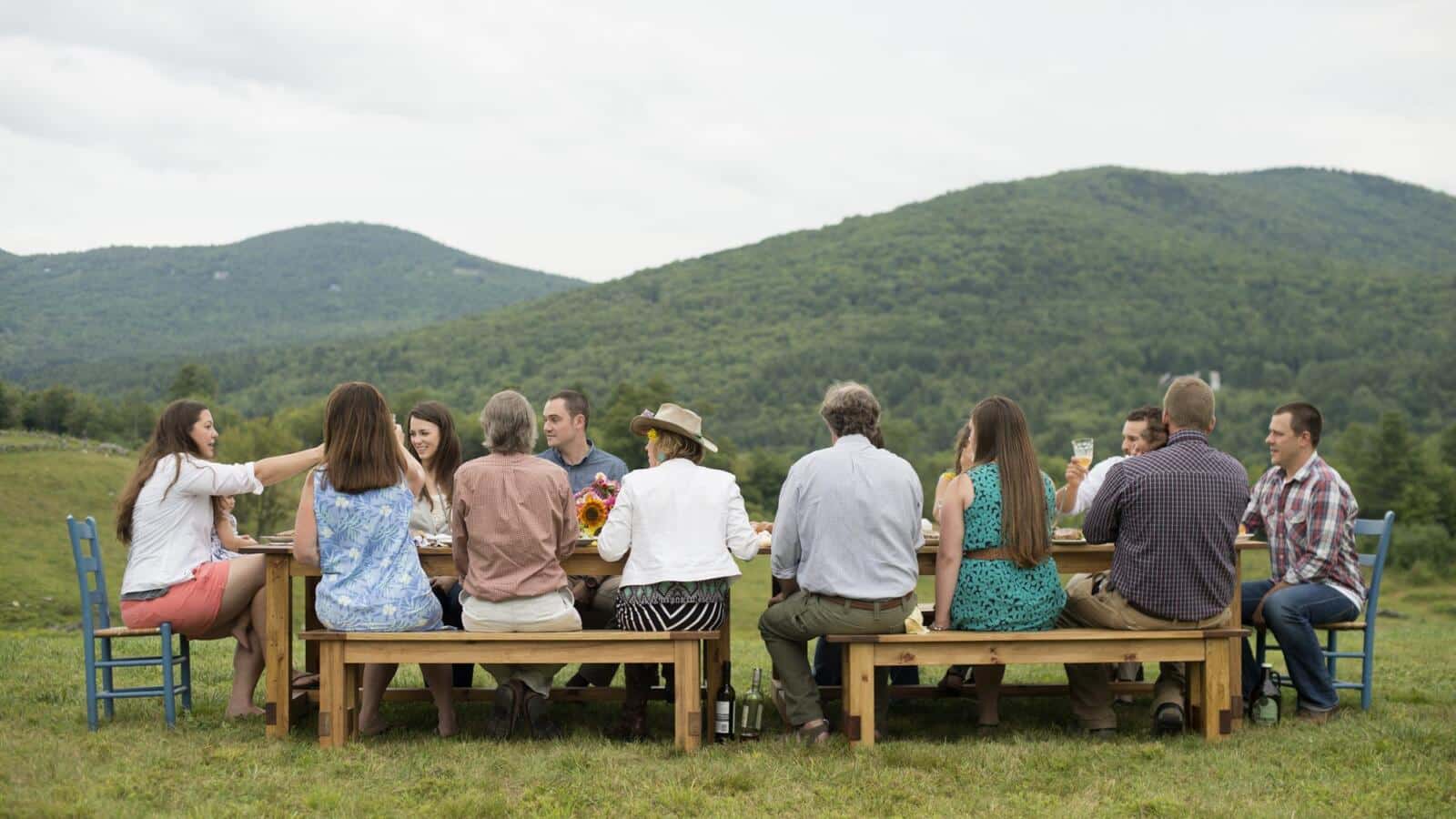 Grupo comiendo en una mesa larga al aire libre con montañas al fondo